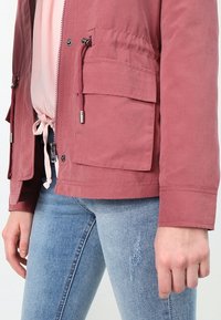 Persona che indossa una giacca color rosa polveroso con zip e tasca a patta, una felpa rosa chiaro sotto e jeans azzurri chiari con piccoli dettagli strappati.