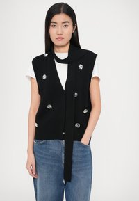 Chaleco negro de punto con escote en V profundo, mangas cortas, adornado con grandes gemas redondas y transparentes. Se ata al frente y se combina con vaqueros azules.