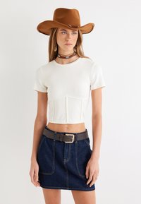 Top corto blanco con mangas cortas, falda de mezclilla de color azul oscuro, cinturón gris, sombrero de fieltro marrón y un gargantilla con acentos en turquesa.