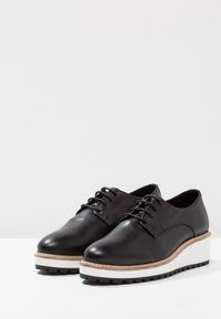 Black leather shoes with a rounded toe, lace-up design, and a chunky white sole featuring a textured black rubber bottom.