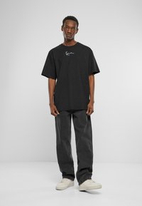 Black oversized cotton T-shirt with short sleeves and a crew neckline, featuring a subtle white logo on the front. Worn with dark gray pants and white sneakers.