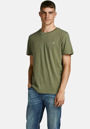 Giovane uomo in piedi, indossa una maglietta a maniche corte verde oliva e jeans blu, con una mano in tasca e sfondo neutro.