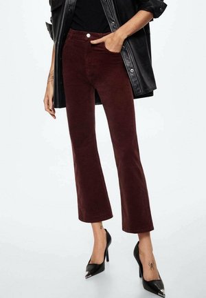 Persoon die cropped bordeauxrode broek, zwarte puntige hoge hakken en een zwart leren jack draagt, met één hand in de zak.