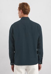 Dunkelgrünes Langarmshirt aus einem strukturierten Stoff, mit klassischem Kragen und Knopfmanschetten, von hinten betrachtet.