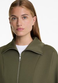 Olivno zelena zip-up jakna z visokim ovratnikom in gladko teksturo. Ima srebrno zadrgo in kontrastno belo notranjo plast.