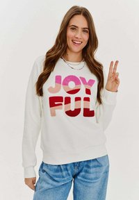 Biela mikina s nápisom "RADOSTNÝ" v jasne ružových a červených plyšových písmenách. Skombinovaná s svetlomodrými džínsami.