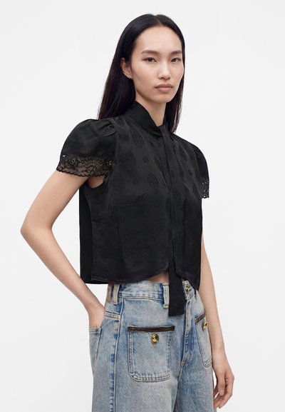 Femme portant un chemisier noir en dentelle à manches courtes avec un col à nouer et un jean bleu clair taille haute avec des poches avant à boutons.