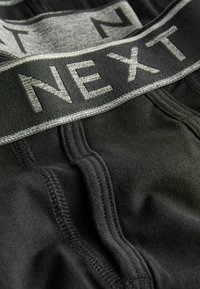 Zwarte katoenen boxershorts met een grijze elastische tailleband waarop "NEXT" in donkere letters is gedrukt. Gladde textuur met contrasterende stikdetails.