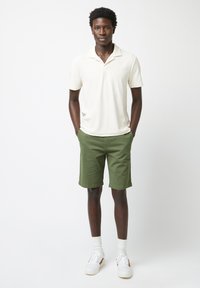 Polo shirt crème avec col, associé à un short vert olive. Baskets blanches avec accents beige et chaussettes blanches. Tenue d'été simple et décontractée.