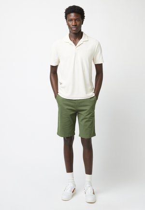 Cremefarbene Polo-Shirt mit Kragen, kombiniert mit olivgrünen Shorts. Weiße Sneakers mit hellbraunen Akzenten und weißen Socken. Einfaches, lässiges Sommeroutfit.