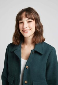 Manteau en laine vert foncé avec un col structuré, des boutons dorés et deux poches avant, porté sur un haut blanc. Tissu texturé.