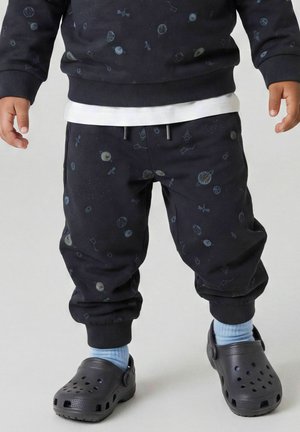 Enfant portant un pantalon noir et un sweat-shirt à motifs sur le thème de l'espace, des chaussettes bleu clair et des chaussures noires sans lacets avec des trous de ventilation.