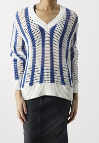 Suéter de punto blanco con rayas azules, escote en forma de V y puños y dobladillo acanalados. Presenta un patrón de tejido abierto. Combinado con una falda negra.