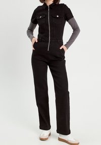 Černý džínový overal s krátkými rukávy, předním zipem, dvěma náprsními kapsami a vysokým pasem, v kombinaci se šedým tričkem s dlouhým rukávem a bílými teniskami.