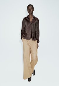 Camicia in raso marrone con bottoni e pantaloni beige a gamba larga. Texture liscia con una vestibilità rilassata; abbinata a scarpe nere.