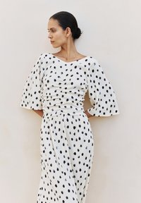 Femme aux cheveux foncés en chignon bas, vêtue d'une robe blanche à manches larges et à pois noirs, debout devant un mur clair uni.