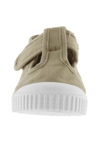 Zapatilla de lona beige con suela de goma blanca, presenta una correa de tipo velcro y un diseño recortado para mayor transpirabilidad. Base plana y texturizada.