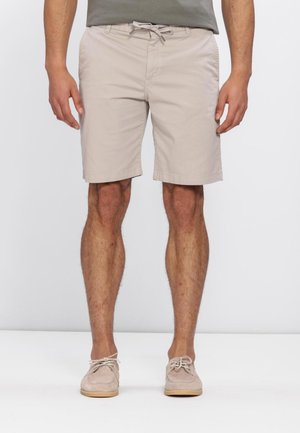 Pantalones cortos beige para hombre hasta la rodilla con cintura con cordón, combinados con zapatos casuales beige claro con cordones, de pie sobre fondo blanco.