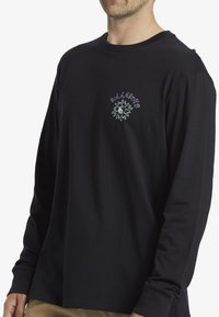 Maglietta nera a maniche lunghe realizzata in tessuto morbido. Presenta una grafica frontale con la scritta "Billabong" e un design del sole in viola e verde.