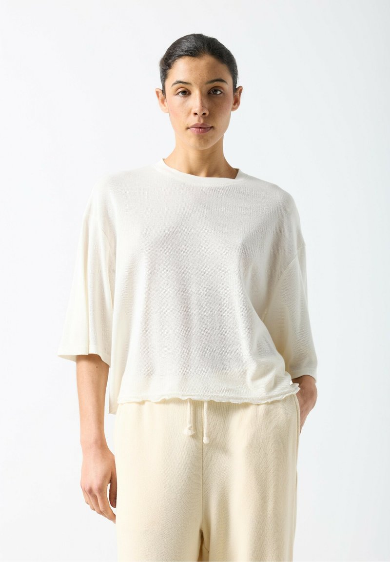 White, loose-fitting short-sleeve top with a textured pattern and a rounded neckline, paired with light-colored, relaxed fit pants.