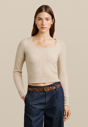 Beige cropped sweater met een vierkante halslijn, geribbelde zoom en lange mouwen, gecombineerd met donkerblauwe high-waisted jeans en een bruine riem.