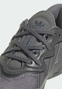 Gray athletic shoe featuring a mesh upper and leather accents, with round laces, textured surface, and a prominent logo on the tongue.