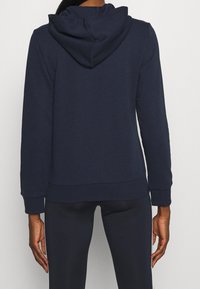 Mörkblå huvtröja med en framficka i känguru-stil, mjukt tyg, ribbade muddarna och luva med snörning, sedd bakifrån.