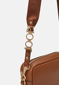Sac bandoulière en cuir marron avec une finition texturée, des accessoires en métal doré et une sangle à motifs avec un logo. Fermeture éclair en haut.