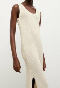 Robe en tricot sans manches couleur crème avec un col rond et une fente sur le côté, portée par une personne à la peau foncée sur un fond clair uni.