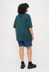 T-shirt oversize verde scuro con maniche corte, abbinato a pantaloni corti in denim. Scarpe chunky nere e calze completano l'outfit.