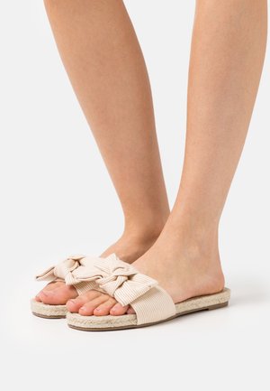 Lichtbeige slide-sandalen met een geknoopte stoffen strikdetail, geweven jute zool en een zachte gestreepte textuur. Het open teenontwerp laat de tenen zien.