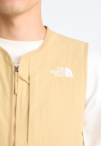 Beige sleeveless vest with a zip closure. Features the white "The North Face" logo on the upper left side. Soft, textured fabric.