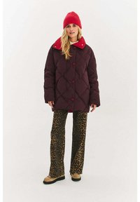 Bordowa pikowana kurtka z czerwonym kołnierzem i guzikami, zestawiona z spodniami w panterkę. Stylizację uzupełnia czerwona dziana czapka.