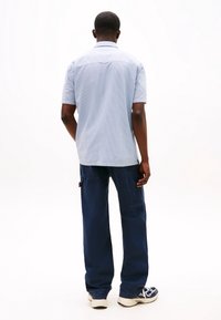 Krátký rukáv, světlá modrá košile s vzorem a límečkem, ve spojení s tmavě modrými kalhotami s širokými nohavicemi a sportovními teniskami, pohled zezadu.