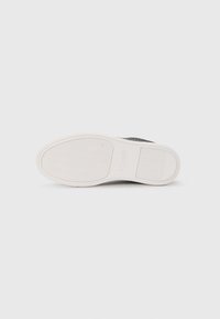 Zwarte textielsneaker met een gestructureerd oppervlak, voorzien van een witte rubberen zool met een golvend patroon en logovermelding op de hak.