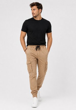 Man met kort donker haar en baard draagt een zwart T-shirt, beige cargobroek met trekkoord, witte sneakers, handen in de zakken, kijkt naar links, staat op een witte achtergrond.
