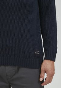 Marineblauer Strickpullover mit strukturiertem Material, gerippten Bündchen und Saum, mit einem kleinen Etikett unten links. Hand, die auf der Seite ruht.