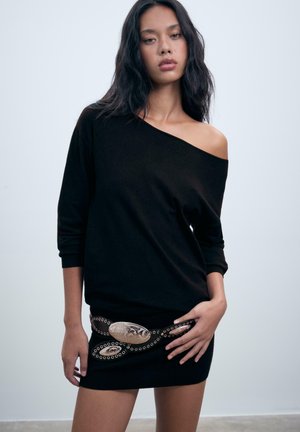Schwarzer schulterfreier Pullover mit langen Ärmeln, kombiniert mit einem eng anliegenden schwarzen Rock mit einem dekorativen metallischen Gürtel.