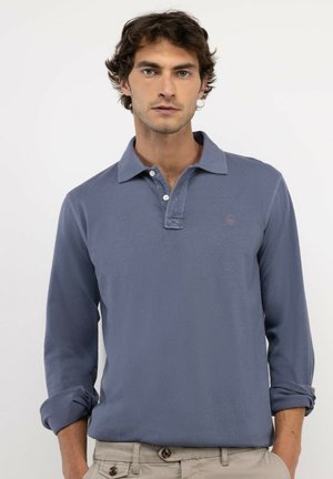Hombre con cabello ondulado que lleva una camiseta polo azul de manga larga con las mangas remangadas y pantalones beige, de pie frente a un fondo liso.