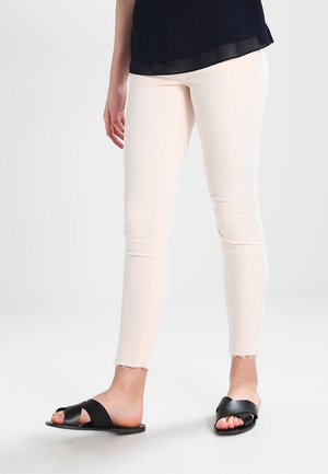 Vrouw die off-white strakke broek met onafgewerkte zomen en zwarte platte sandalen met gekruiste bandjes draagt, staand op een lichtgrijze vloer.