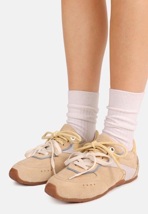 Μπεζ σουέτ αθλητικά παπούτσια με ανοιχτό μπλε λεπτομέρειες, στρογγυλή μύτη και κρέμ κορδόνια. Φορεμένα με λευκές ριγωτές κάλτσες, με καφέ λαστιχένια σόλα.