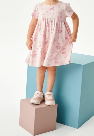 Robe en coton rose ornée d'un motif répétitif Peppa Pig, manches courtes et taille froncée. Un enfant se tient debout sur des blocs colorés, chaussé de baskets rose clair.