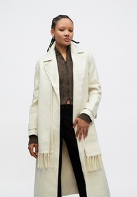 Biely vlnený kabát s trsťovým detailom, výstrihom s výrezom a bočnými vreckami, vrstvený cez hnedý rebrovaný sveter a čierne nohavice. Hladká textúra.