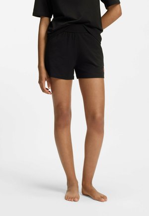 Personne portant un short noir à taille élastique et un t-shirt noir, debout pieds nus sur un fond blanc uni.