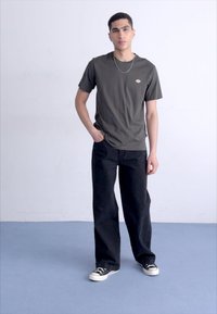 Tricou verde măsliniu cu un logo mic, purtat cu blugi negri lejeri și adidași alb-negru, stând pe un podea de culoare albastră deschisă.