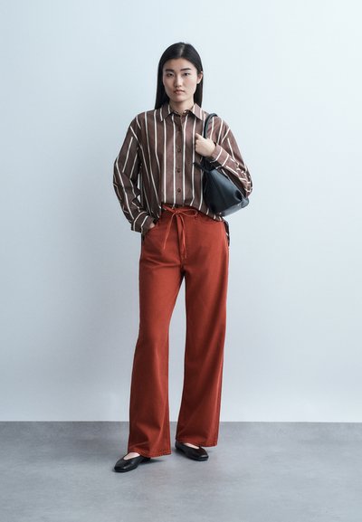 Γυναίκα όρθια με καφέ ριγέ πουκάμισο, φαρδιά παντελόνια σε σκουριά απόχρωση, μαύρες μπαλαρίνες και μαύρη τσάντα ώμου σε ανοιχτό γκρι φόντο.