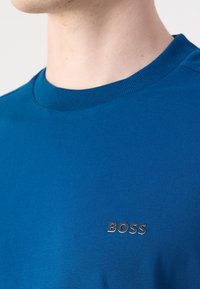 Modrá bavlněná tričko s žebrovaným kulatým výstřihem a stříbrným logem "BOSS" na levém prsu. Hladká textura.