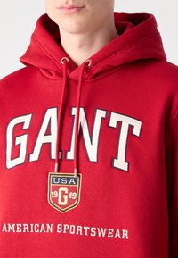 Sudadera de algodón roja con el texto "G.A.N.T" en blanco y azul, un parche de logo amarillo y azul, y las palabras "AMERICAN SPORTSWEAR" debajo.