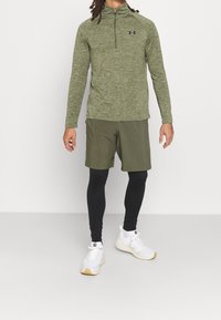 Olivgrön zip-up långärmad tröja i lättviktsmaterial, matchad med olivgröna shorts, svarta kompressionsbyxor och vita träningsskor.