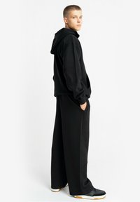 Młoda osoba z krótkimi włosami, ubrana w czarną bluzę z kapturem i szerokie czarne spodnie, stojąca bokiem z rękami w kieszeniach na białym tle.
